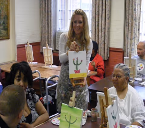 A group of people sitting around a table in a room, participating in a painting activity. A woman standing in the center is holding up a painting of a cactus in a pink pot. Other participants have similar cactus paintings on small easels in front of them. The room has large windows with curtains and wooden chairs.