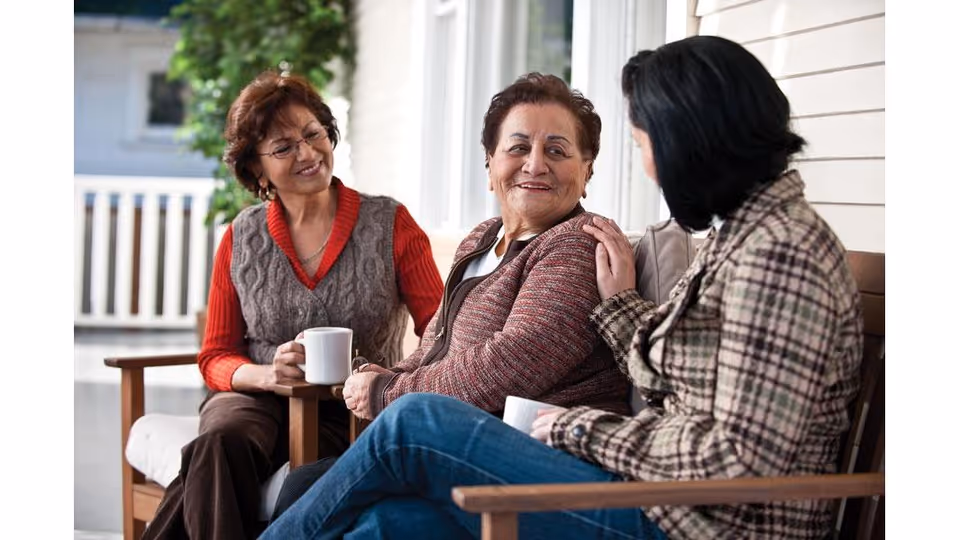 Three women sitting on a porch bench, engaged in conversation and holding white mugs. One woman is gently touching the shoulder of another, and they all appear to be enjoying each other's company in an outdoor setting with a white railing and greenery in the background.
