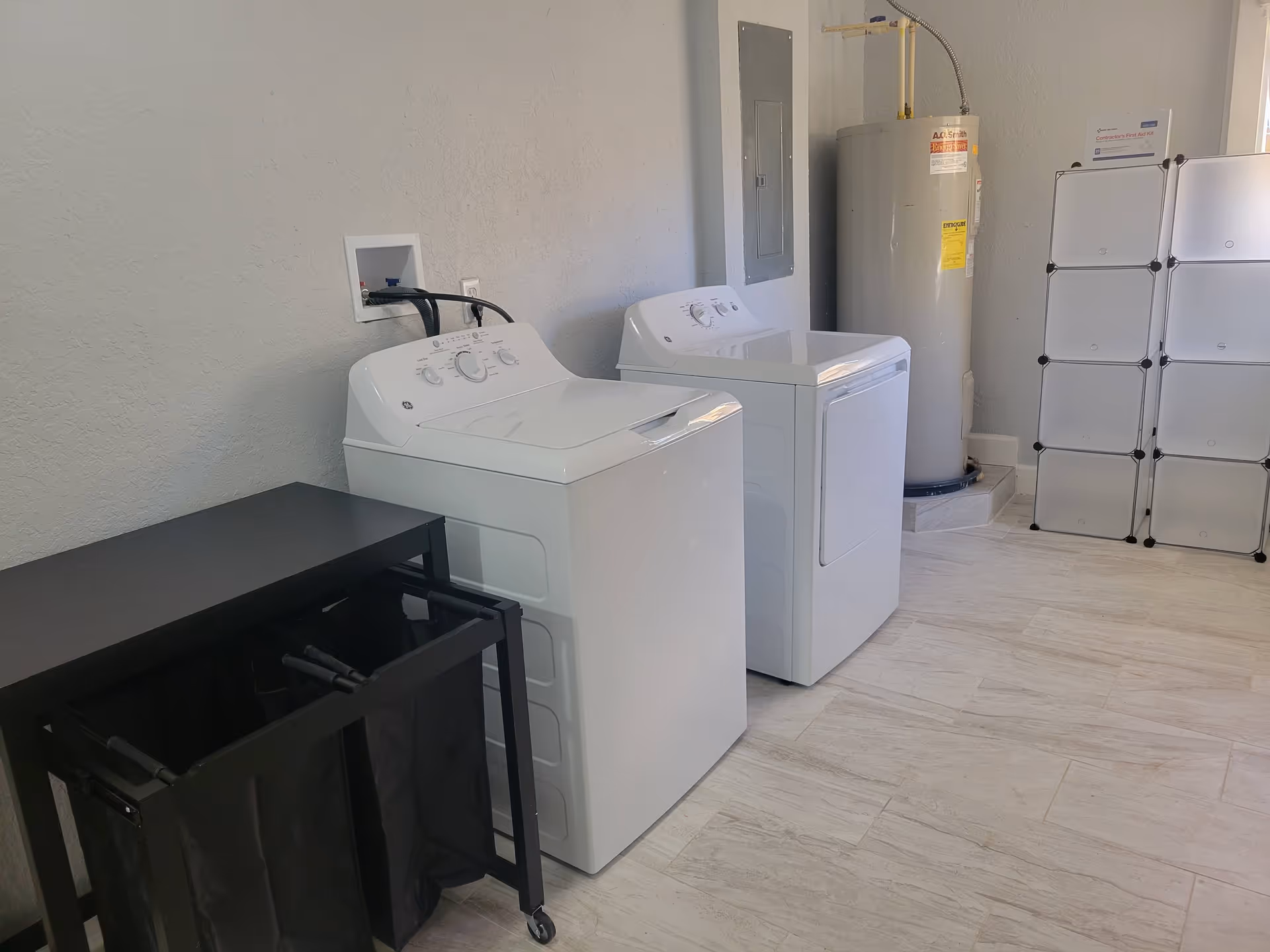 A laundry room with a white washing machine and dryer side by side, a black laundry sorter with two compartments on wheels, a water heater in the corner, and a white modular storage unit against the wall.