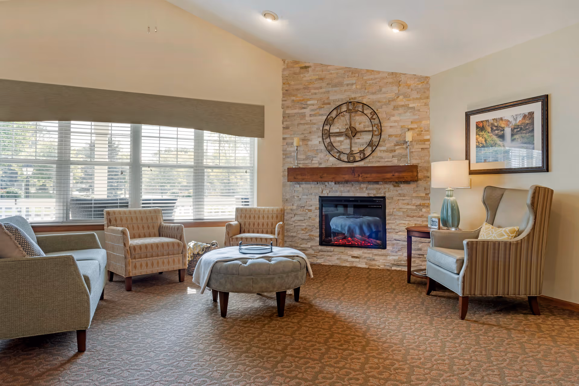 A cozy living room area with a stone fireplace featuring a large round clock above the mantel. The room has a large window with blinds, two patterned armchairs, a gray sofa, a round ottoman with a blanket, a side table with a lamp, and a framed picture on the wall.