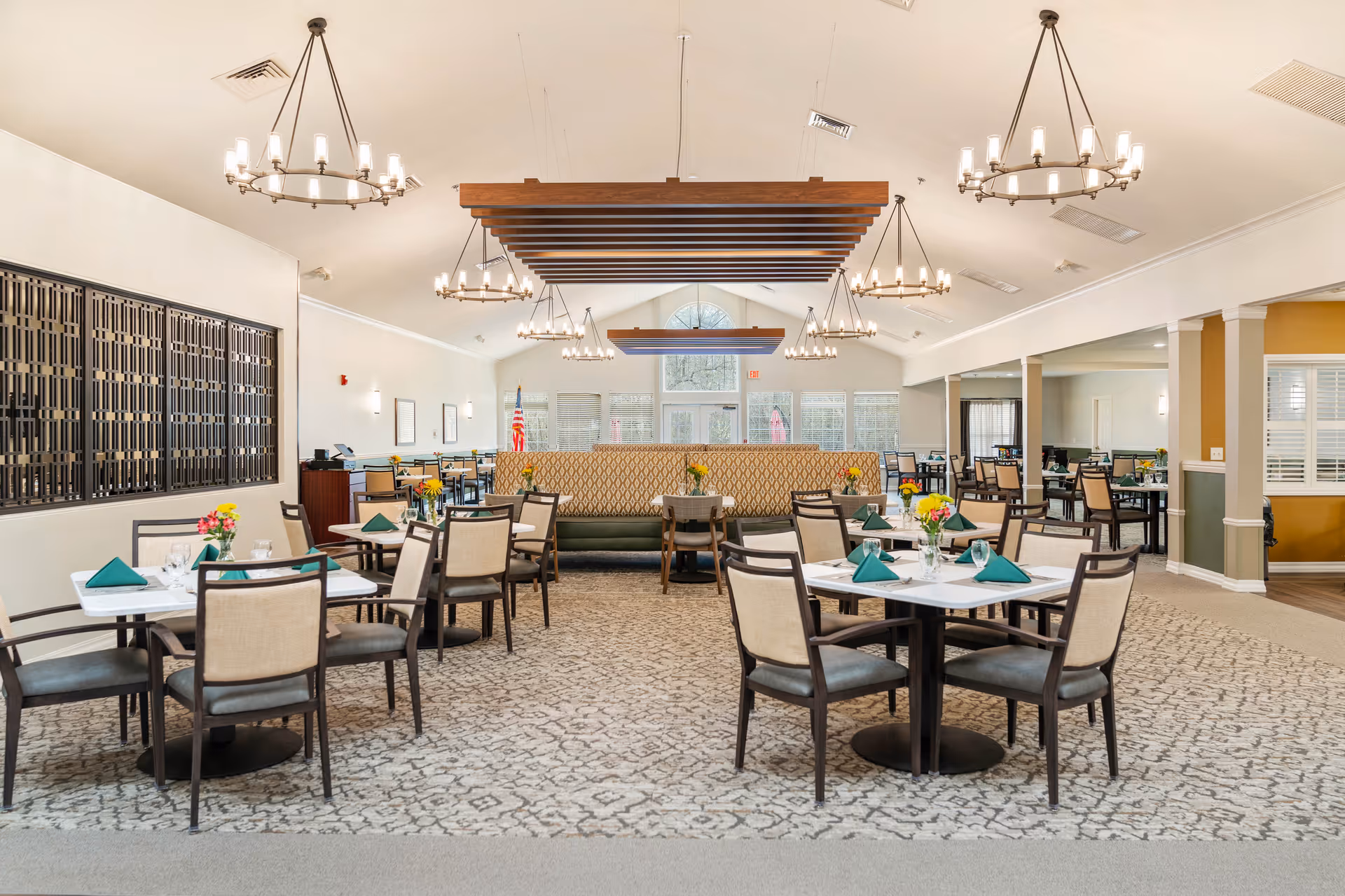 Spacious dining room with multiple set tables, chandeliers, and a central banquette beneath wooden ceiling beams.