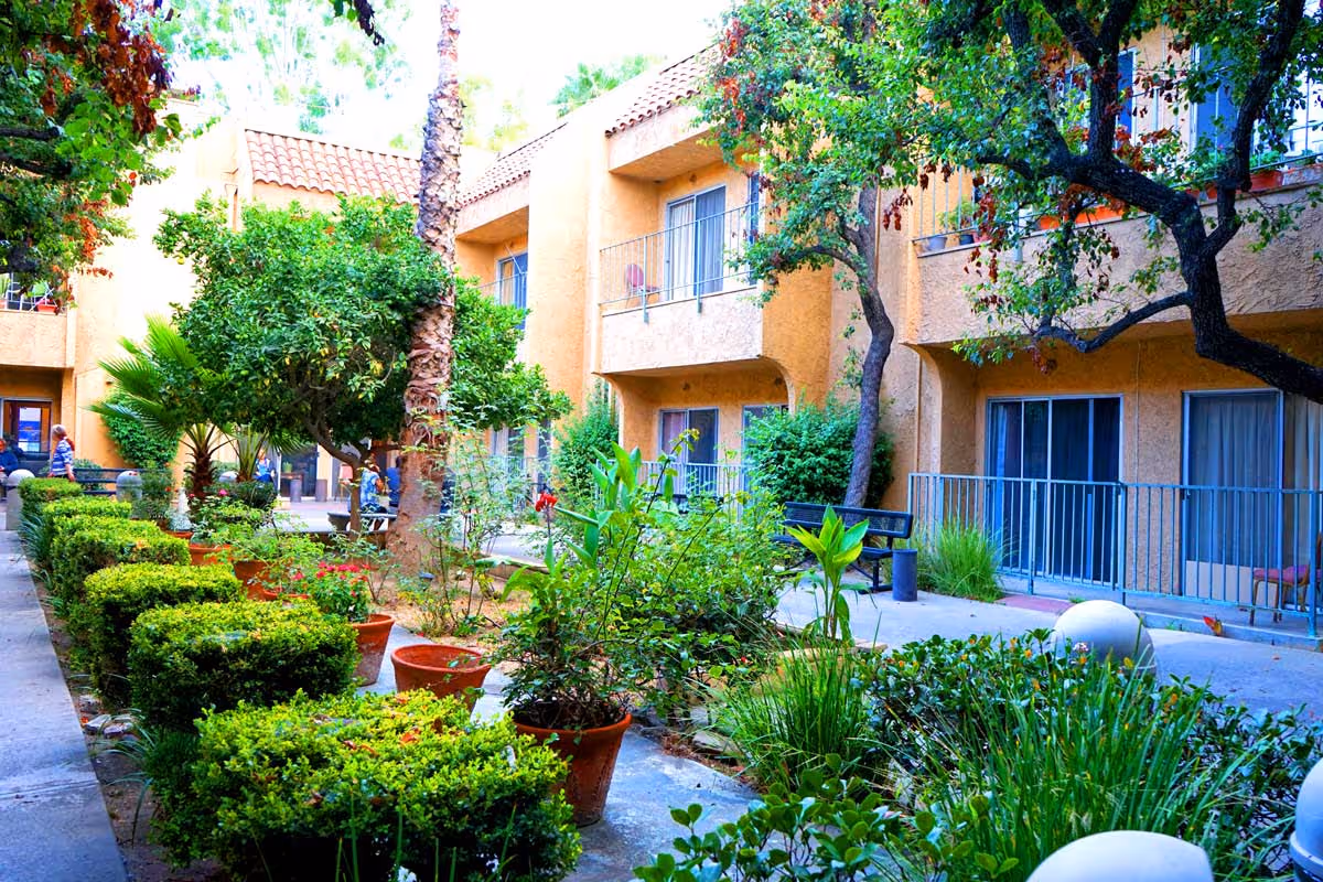 Outdoor courtyard area of a retirement home with lush green plants, potted flowers, trimmed bushes, and trees. The courtyard is surrounded by a two-story building with balconies and sliding glass doors. Benches are placed along the walkway for seating.