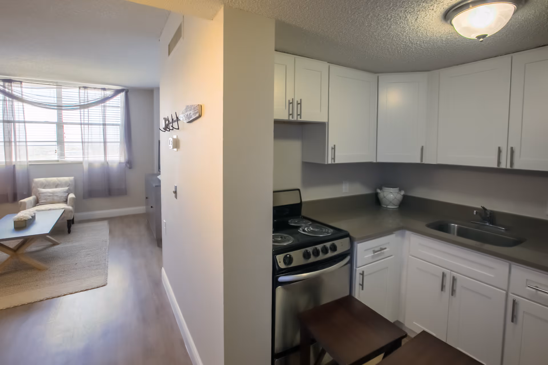 Open-plan apartment showing a small kitchen with white cabinets, a stainless steel stove and sink, and a living area with an armchair and coffee table.