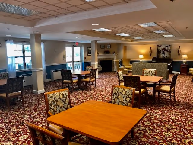 A spacious senior living common area with multiple wooden tables and floral upholstered chairs arranged on a patterned carpet. The room features large windows with white curtains, a fireplace, and soft lighting from table lamps and wall sconces, creating a warm and inviting atmosphere.