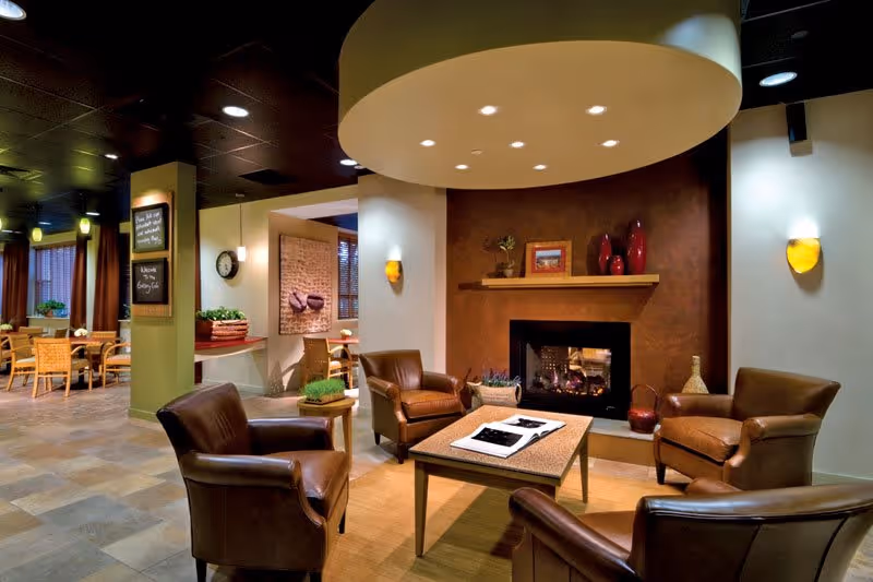 A cozy lounge area with four brown leather armchairs arranged around a rectangular coffee table with a book on it. The room features a modern fireplace with decorative items on the mantel, warm lighting, and a circular ceiling fixture above the seating area. In the background, there is a dining area with wooden chairs and tables, and a chalkboard menu on a green pillar.