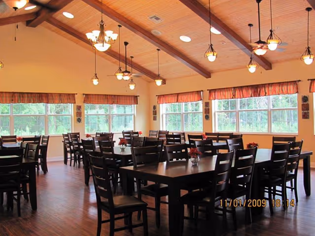 A bright dining room with multiple dark wood tables and chairs, a vaulted wood ceiling with hanging lights, and large windows along the walls.
