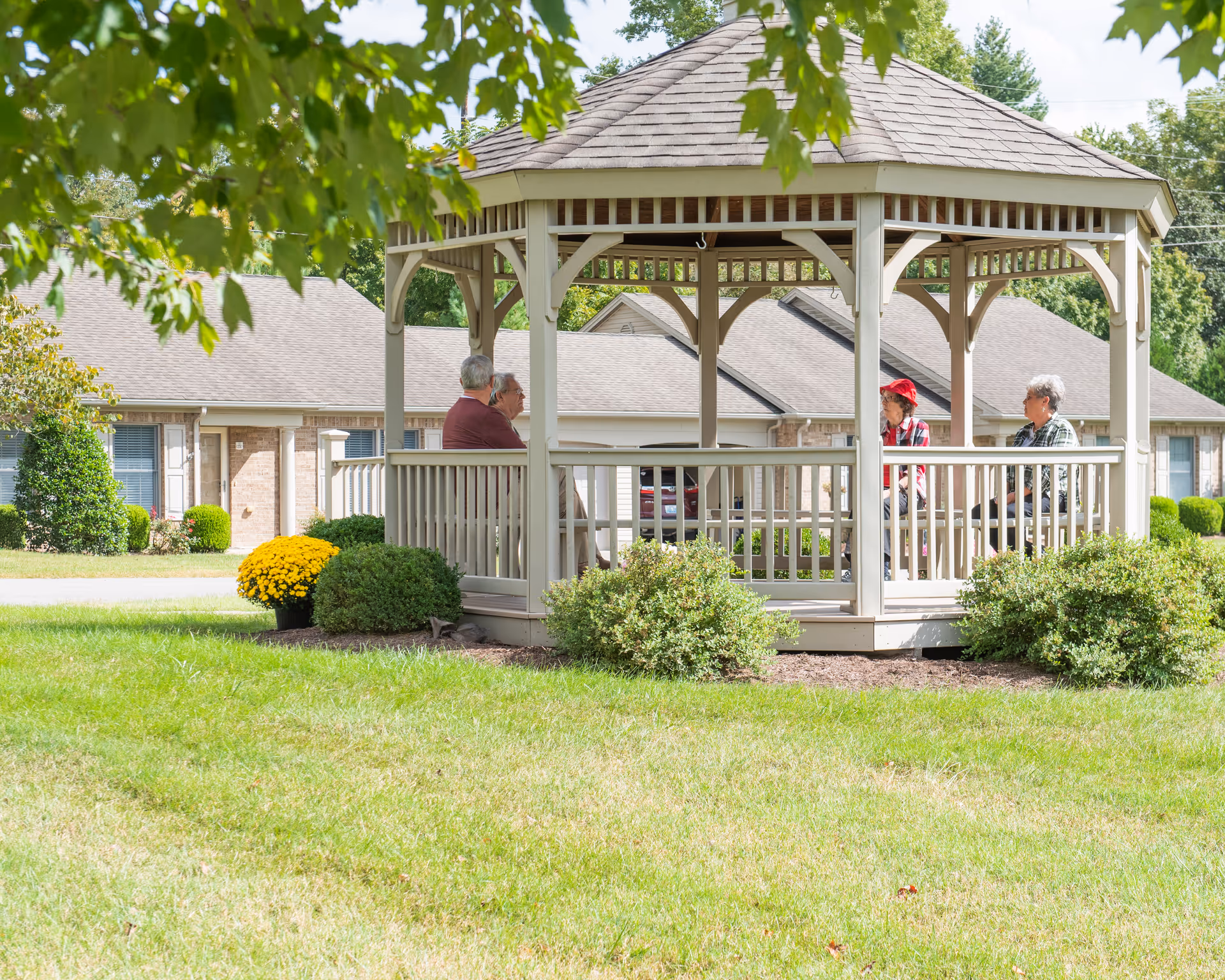 Four elderly people sitting and talking inside a wooden gazebo in a grassy outdoor area with bushes and yellow flowers, with residential buildings in the background.