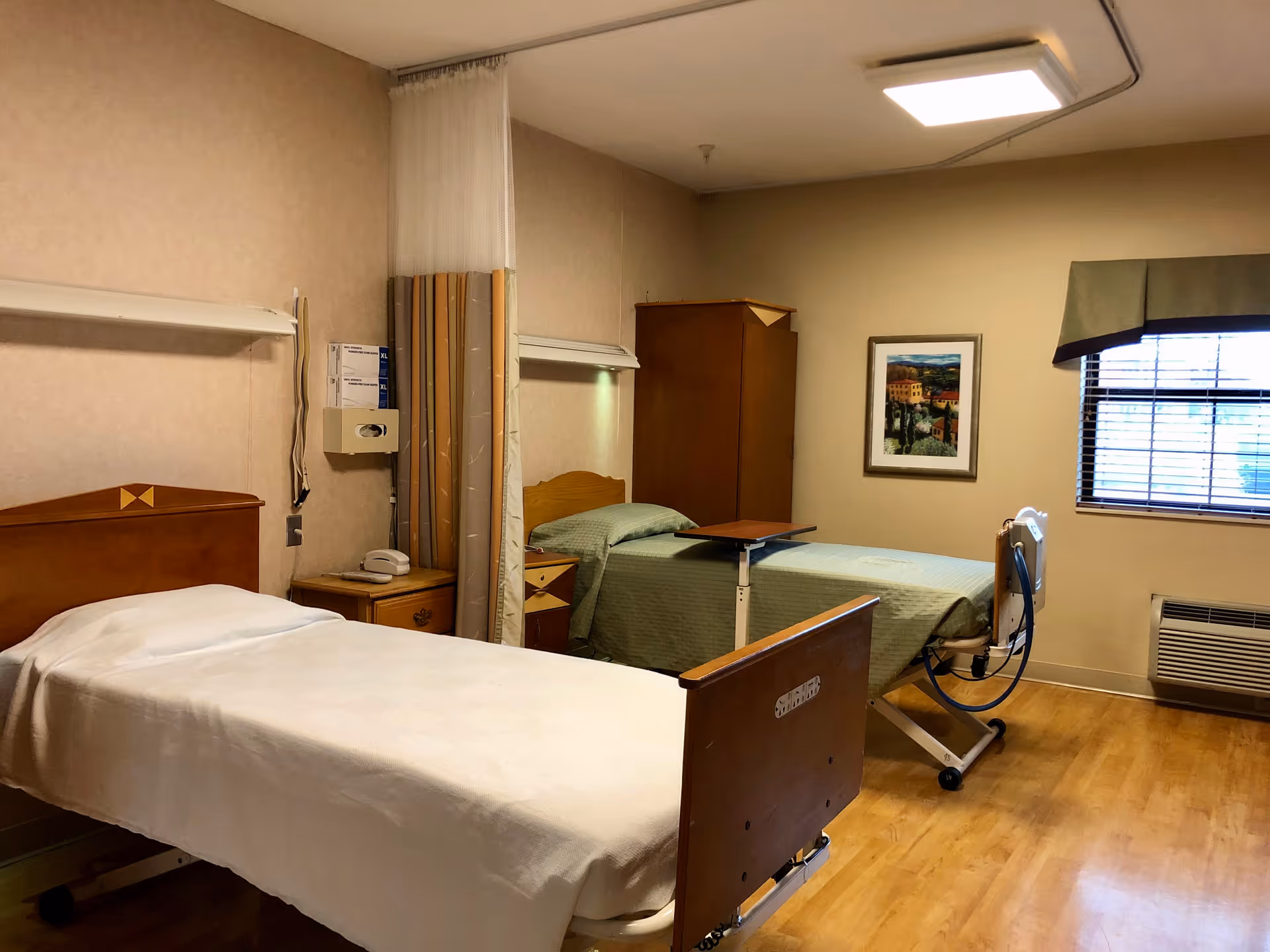 Two hospital-style beds in a senior living facility room with wooden floors, bedside furniture, a privacy curtain, and a window.