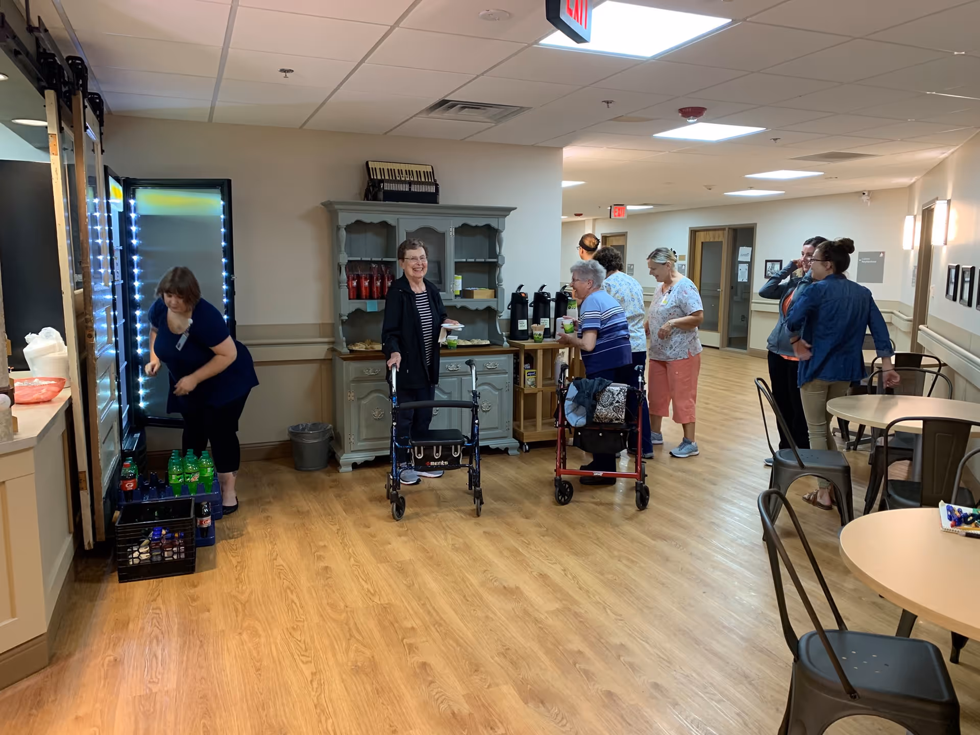 A group of elderly people and caregivers gathered in a common area of a senior living facility. Two elderly women with walkers are near a cabinet with coffee dispensers and snacks. Other people are standing and conversing near tables and chairs. The room has wood flooring, beige walls, and ceiling lights.
