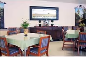 Dining room with square tables covered in light green tablecloths, wooden chairs, a sideboard with a mirror and decorative plants and artwork.