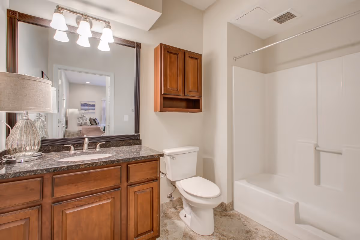 A clean and well-lit bathroom featuring a wooden vanity with a granite countertop and an undermount sink. Above the sink is a large mirror with a dark wooden frame and a light fixture with four bulbs. To the right of the vanity is a white toilet and a wooden wall cabinet mounted above it. Further right is a white bathtub with a shower curtain rod installed above it. The floor is tiled with a beige pattern.