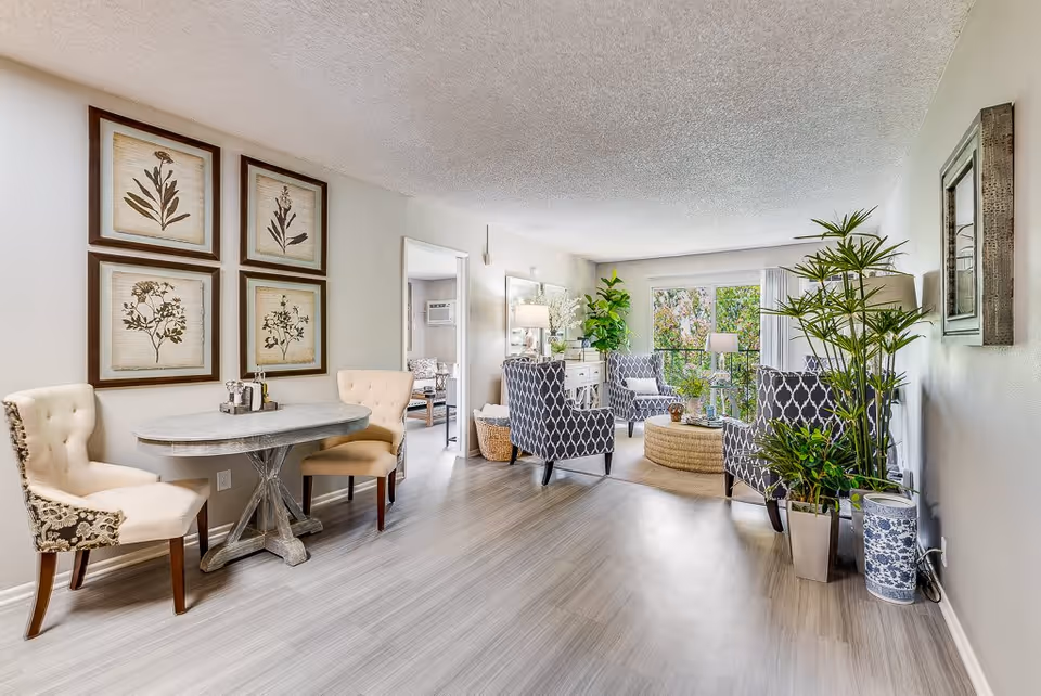 A bright and spacious living room area with light-colored wooden flooring. On the left side, there is a small round table with two upholstered chairs. Above the table, four framed botanical prints are hung on the wall. The living area features three patterned armchairs arranged around a round woven coffee table. There are several potted plants, including a tall one near the right wall and another near the window. A large window at the back lets in natural light, and a doorway leads to another room with more seating visible.