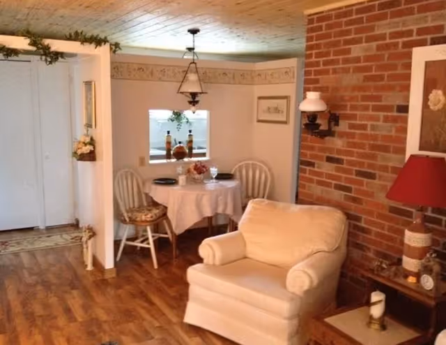Cozy living room and dining nook with an upholstered armchair, brick accent wall, and a small round table set for two.