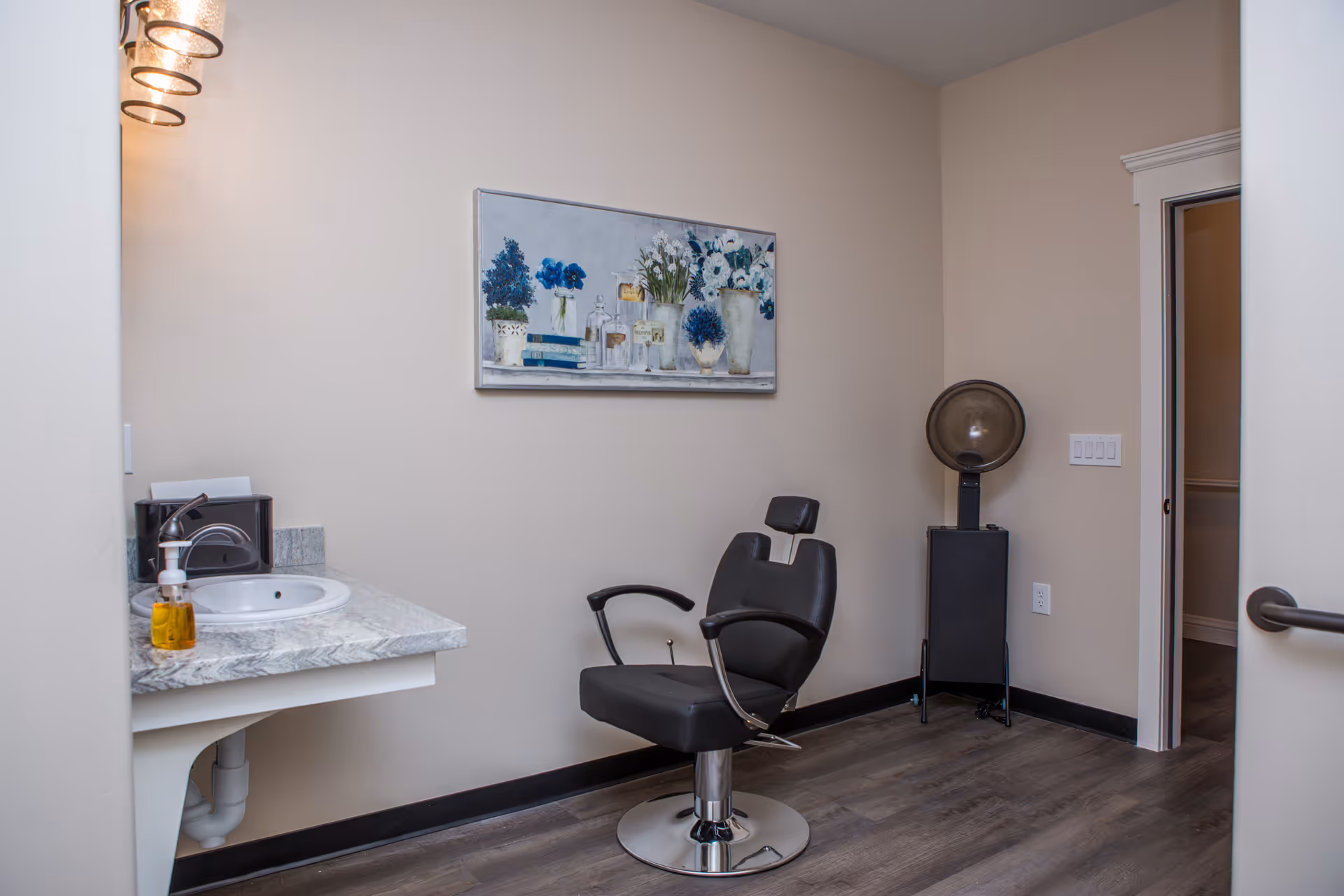 A small room with a black salon chair and a hair dryer hood in the corner. There is a countertop with a sink, soap dispenser, and tissue box on the left side. A painting of blue and white flowers hangs on the beige wall above the chair. The floor is wood, and the room has a clean, simple design.