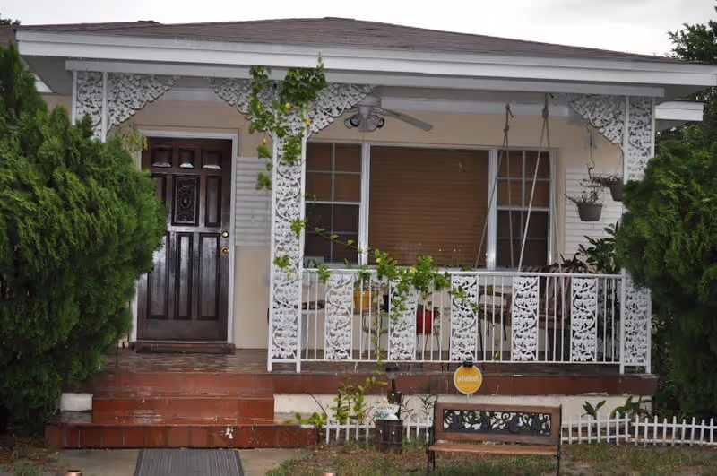 Front porch of a house with a dark wooden door, white decorative railing and trim, a porch swing, potted plants, and two large green bushes on either side. There is a small bench and a security sign in the front yard.