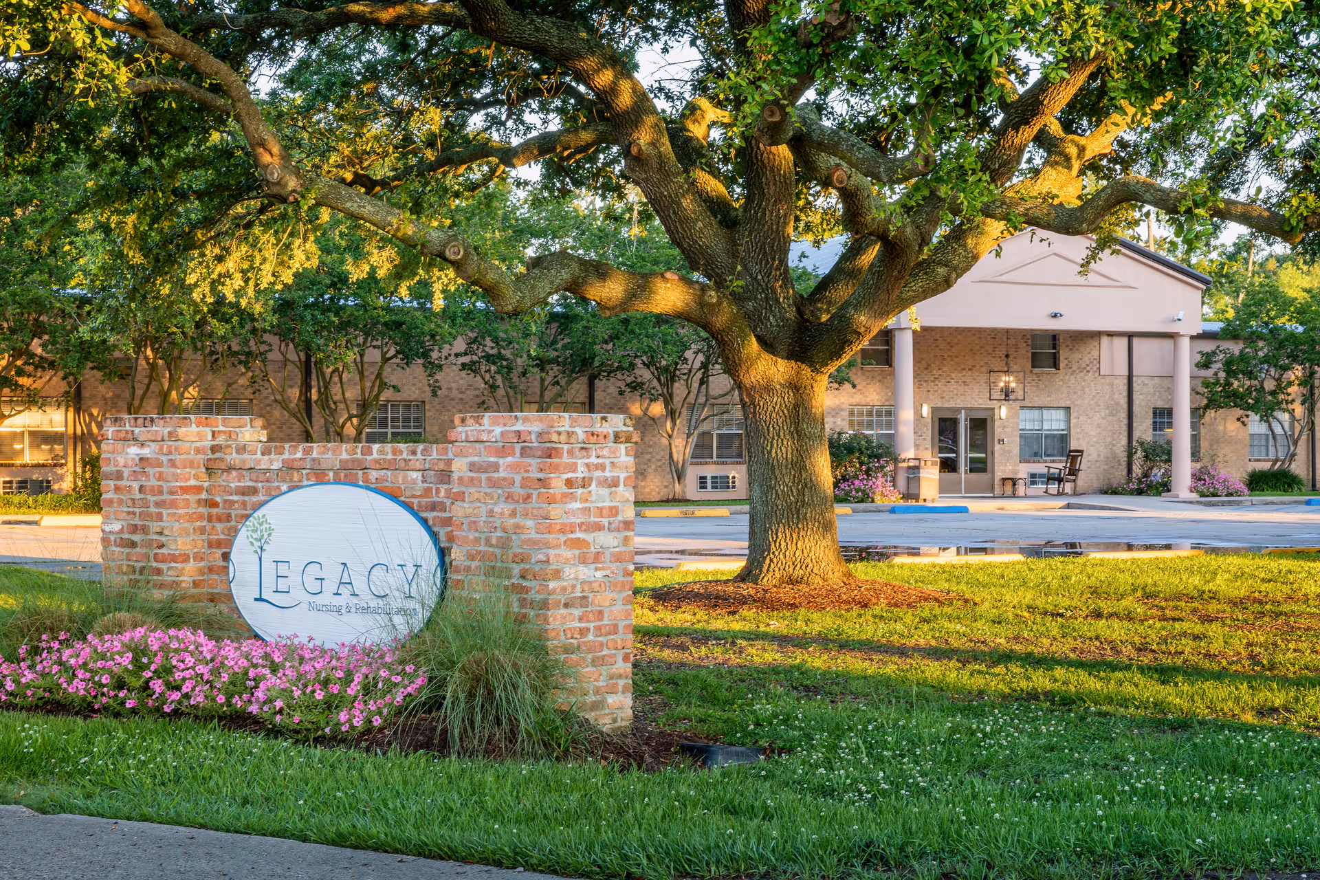 Exterior view of Legacy Nursing and Rehabilitation of Port Allen facility with a large tree, a brick sign with the facility name, green grass, and flowers in the foreground.