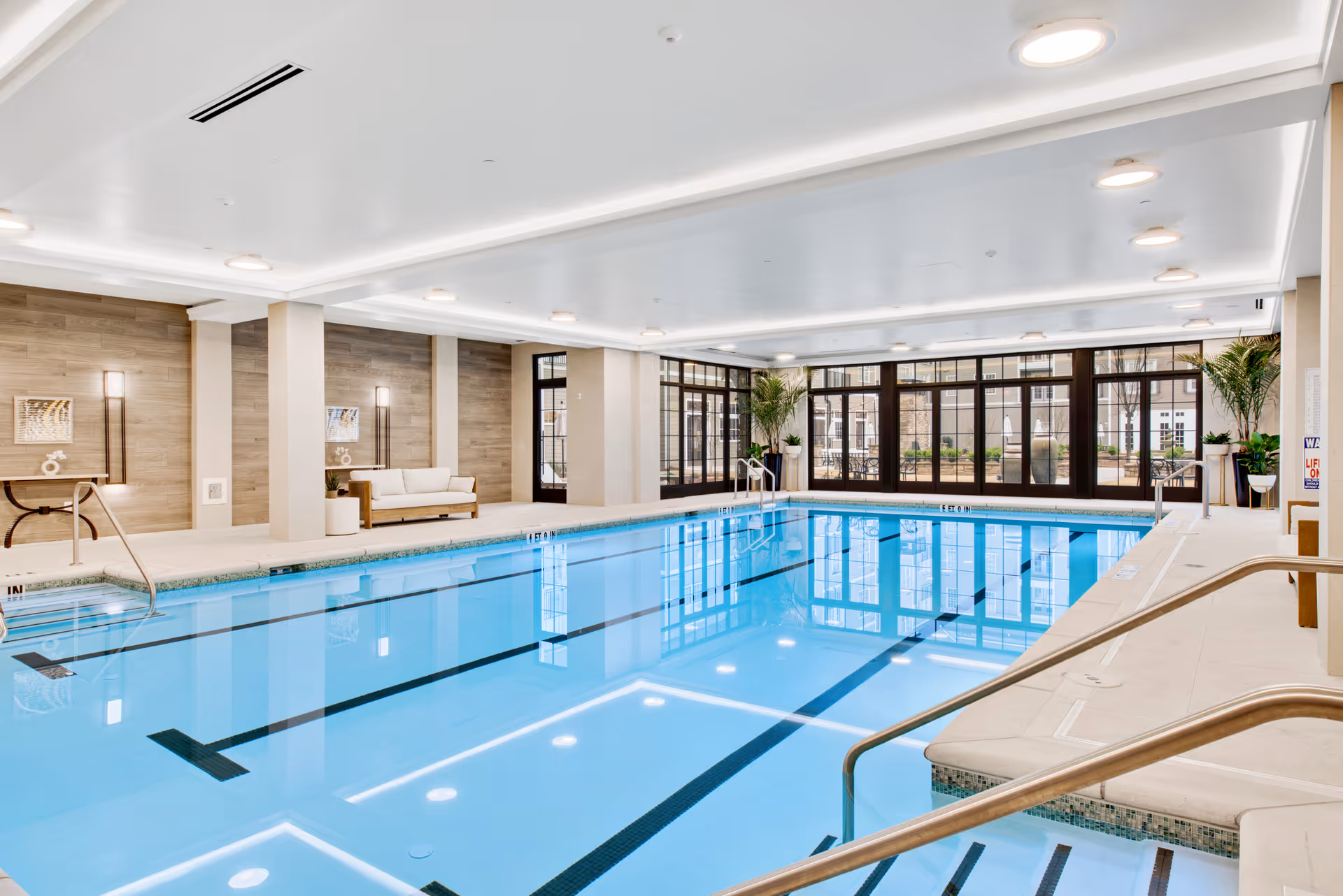 Spacious indoor swimming pool with lounge seating, large windows, and handrails in a bright facility room.