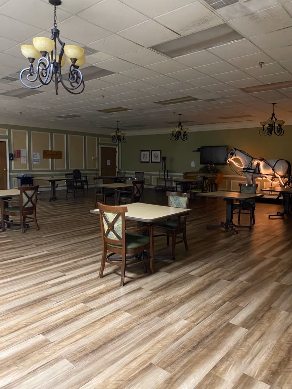 A spacious dining room with multiple tables and chairs arranged on a wooden floor. The walls are painted green with white trim, and several chandeliers hang from the ceiling. In the background, there is a decorative illuminated horse sculpture and a wall-mounted television.