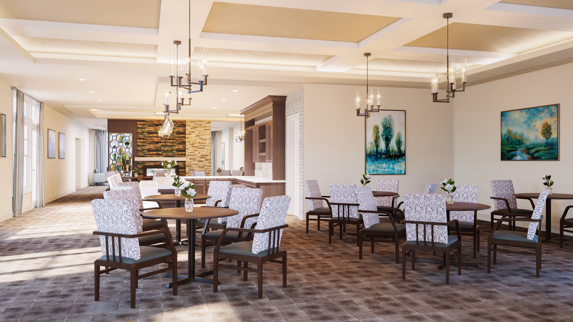 A spacious dining area in a senior living facility with multiple round tables surrounded by cushioned chairs. Each table has a small vase with flowers. The room features large windows allowing natural light, modern chandeliers hanging from a coffered ceiling, and artwork on the walls. The floor is tiled with a patterned design, and there is a stone accent wall in the background.
