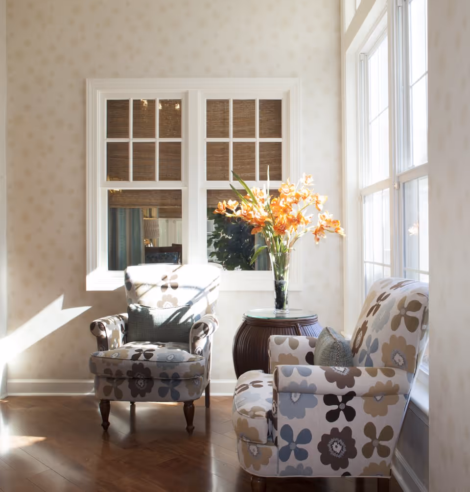 A bright sitting area with two floral patterned armchairs facing each other, a small round wooden table between them holding a vase with orange flowers, and large windows letting in natural light.