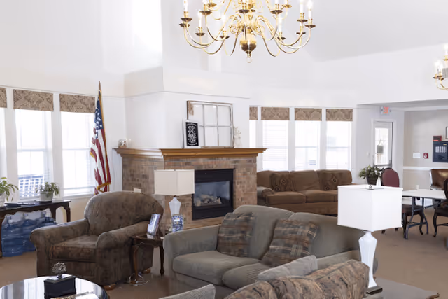 A bright and spacious living room area with multiple upholstered sofas and armchairs arranged around a brick fireplace. The room features large windows with patterned valances, a chandelier hanging from the ceiling, an American flag in the corner, and several table lamps. There are also some plants and a table with chairs in the background.