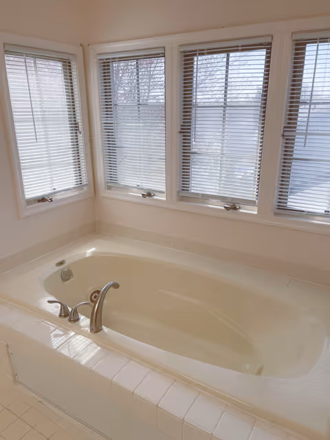 A built-in white soaking bathtub beneath a row of windows with closed blinds in a bright bathroom.