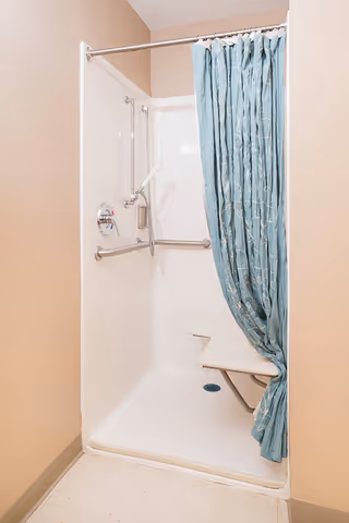 A white walk-in shower with a blue patterned shower curtain partially drawn to the side. The shower features a fold-down seat, grab bars for safety, and a handheld showerhead. The walls are beige and the floor outside the shower is light-colored.
