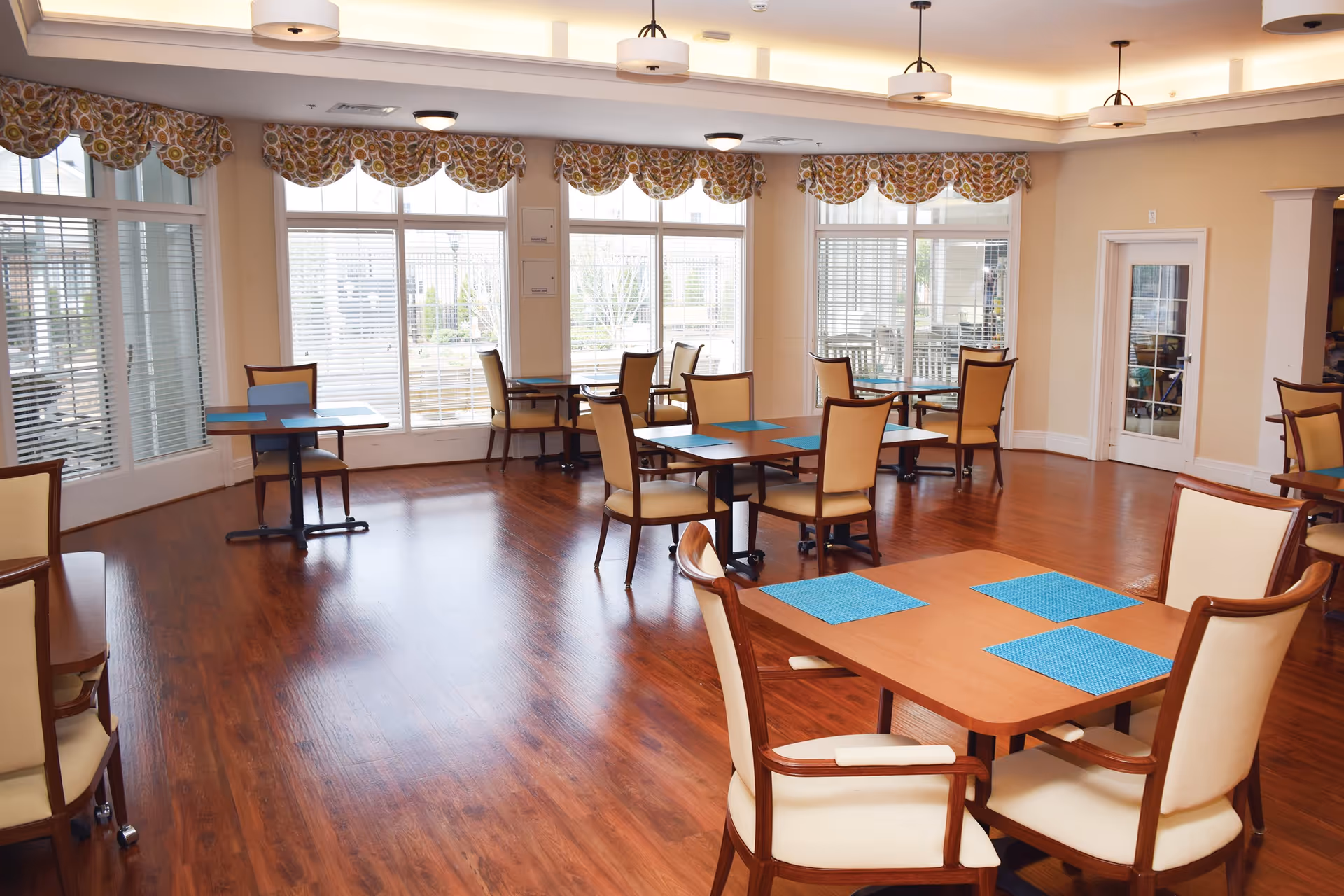 A bright dining room with wooden floors, several tables with blue placemats, and beige cushioned chairs. Large windows with patterned valances allow natural light to fill the room.
