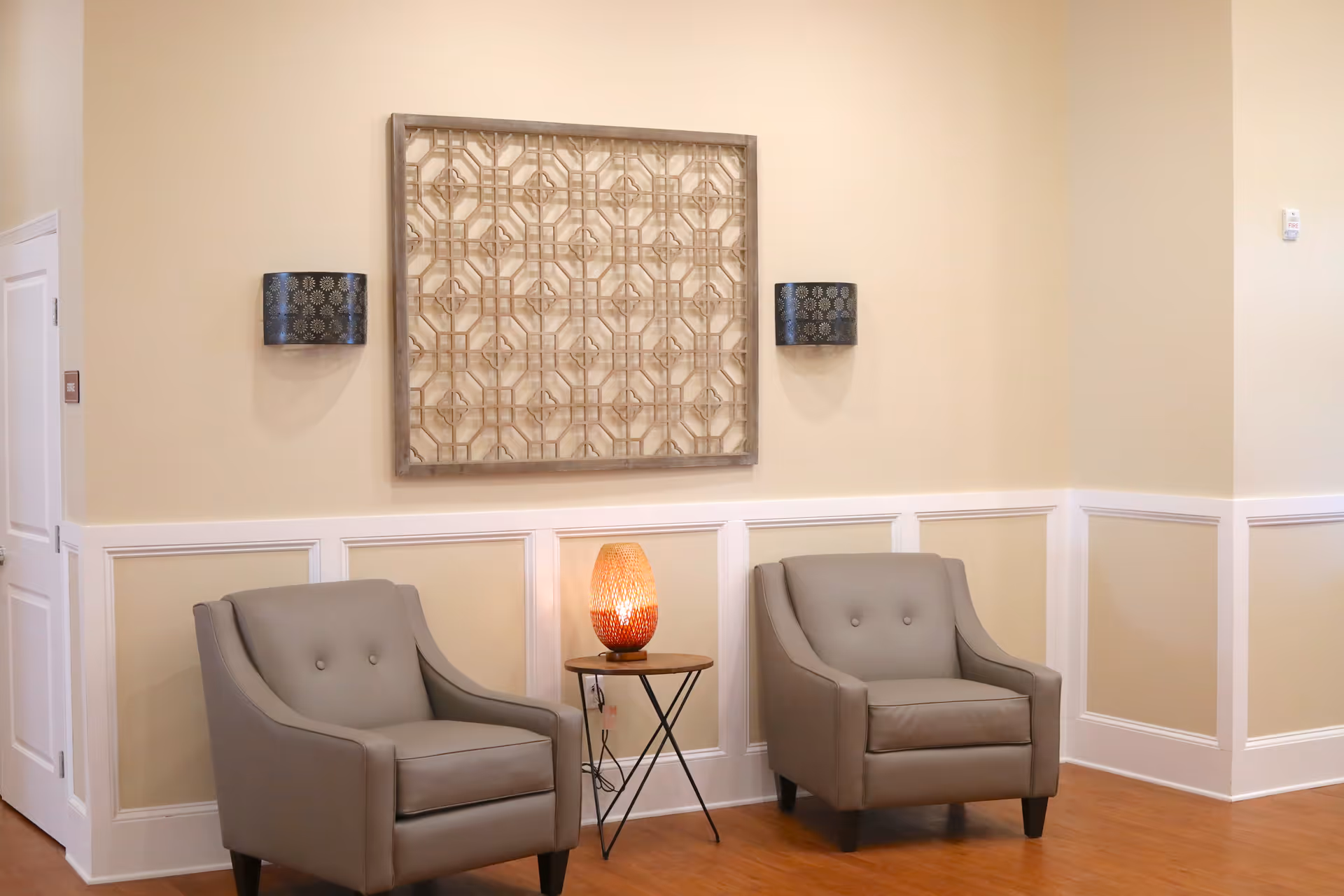 A cozy seating area with two gray armchairs separated by a small round wooden table holding a decorative lamp. The walls are painted beige with white wainscoting, and a large decorative wooden wall art piece hangs above the table, flanked by two black patterned wall sconces.