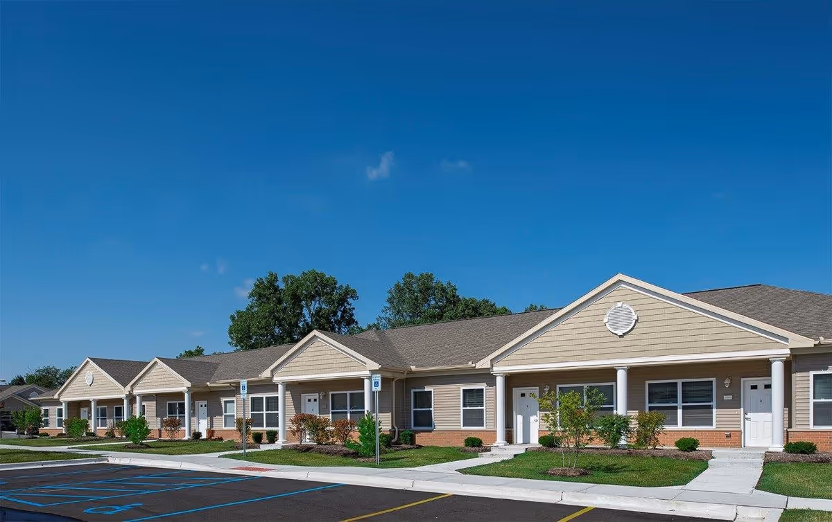 Exterior view of a single-story residential building with multiple entrances, beige siding, white trim, and a gray roof under a clear blue sky. There are small landscaped areas with bushes and trees in front, and a parking lot with marked spaces including handicapped spots.