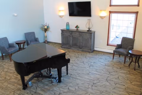 Bright common room with a black grand piano, several chairs and side tables, a wooden cabinet with a mounted TV, and windows.