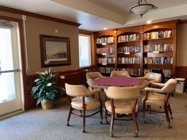 A cozy room with a round table covered with a purple felt top surrounded by six beige cushioned chairs on wheels. Behind the table is a large wooden bookshelf filled with books. To the left, there is a potted plant and a framed picture on the wall next to a glass door and window letting in natural light. The room has beige walls with wooden paneling and a ceiling light fixture.