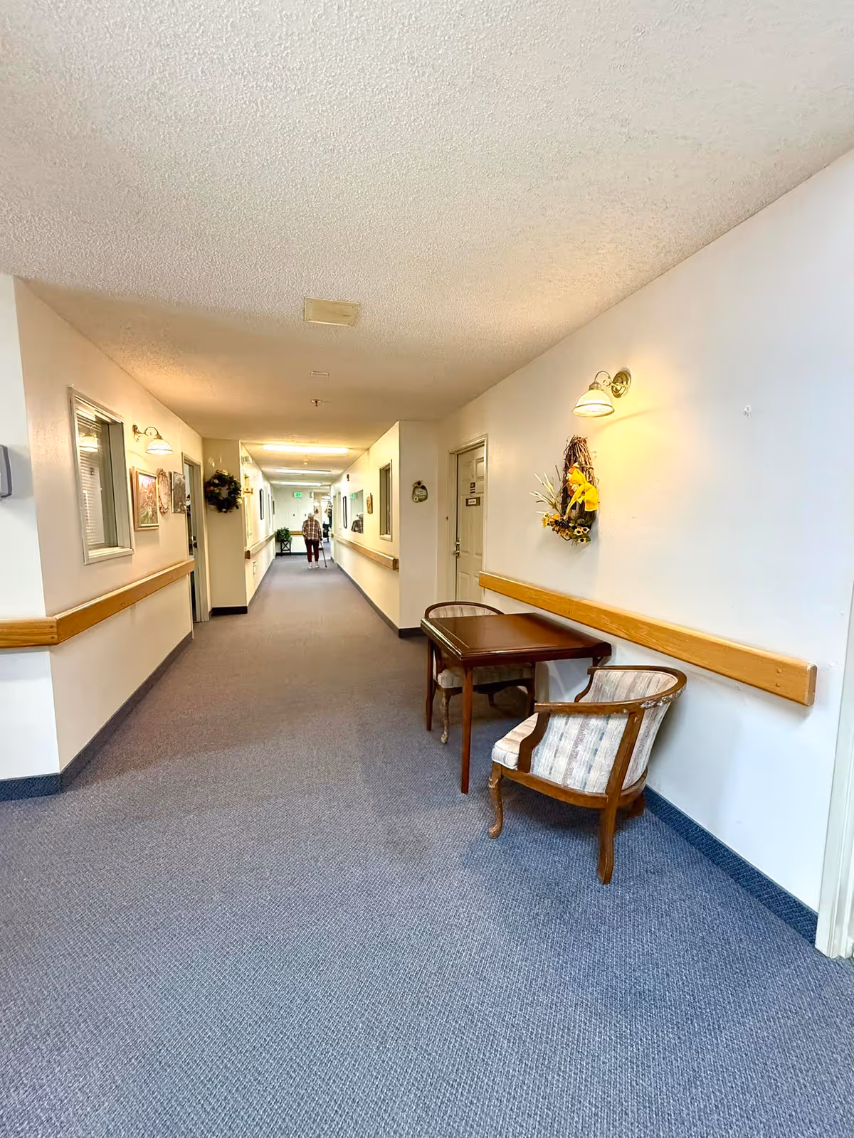A long, carpeted hallway in a retirement center with white walls and wooden handrails. There are framed pictures and decorative wreaths on the walls. A small table with two upholstered chairs is placed along the right wall. A person is walking down the hallway in the distance.
