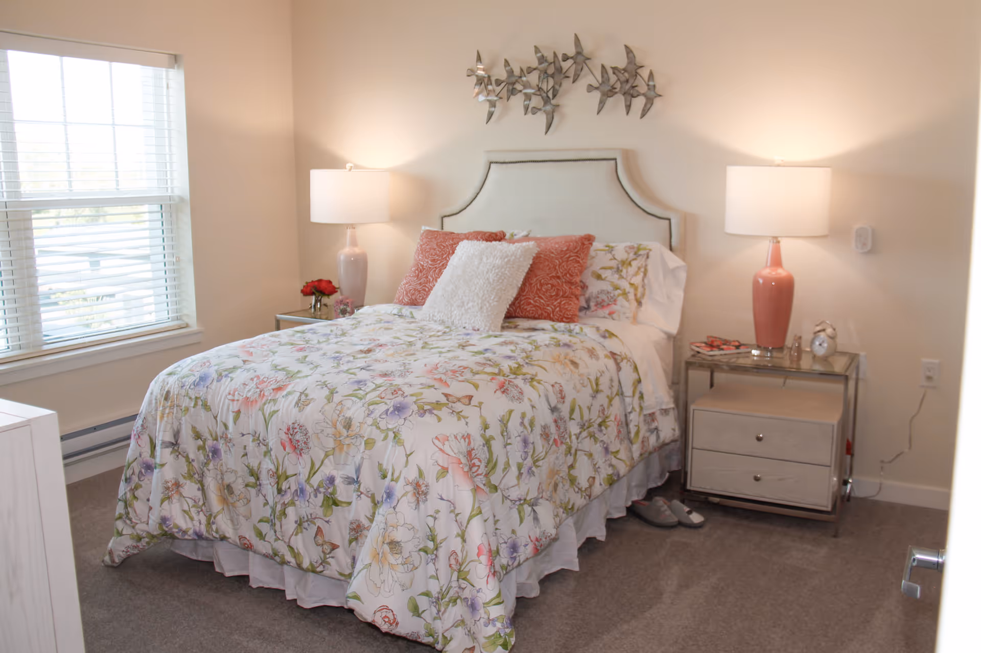 Bright bedroom featuring a floral-covered bed with a headboard, two bedside tables with lamps, and a window to the left.