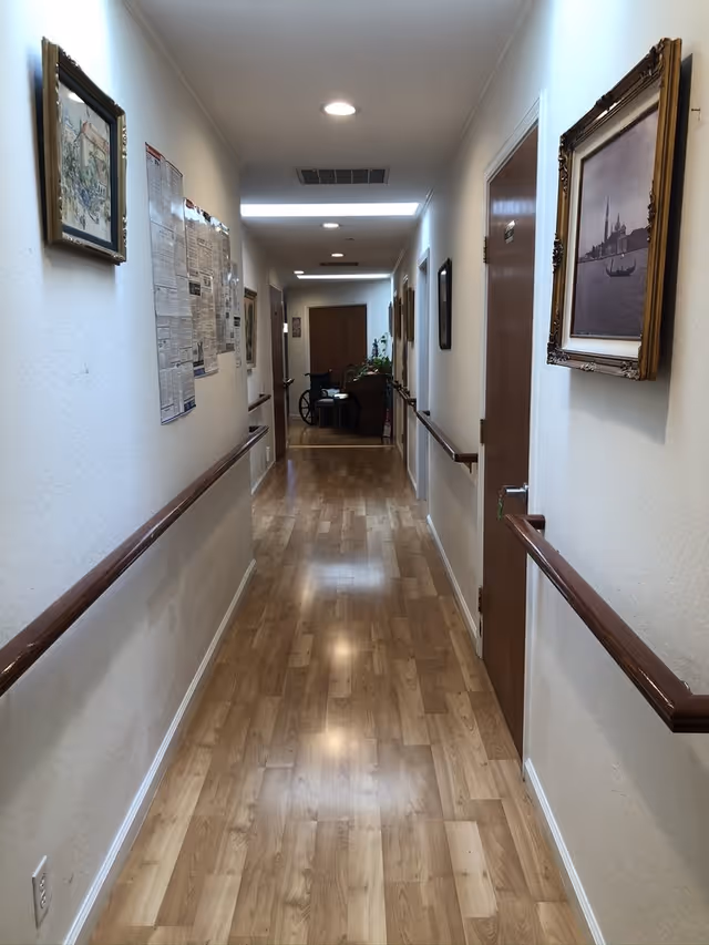Well-lit interior hallway with wood flooring, handrails on both sides, framed artwork, and a wheelchair at the far end.