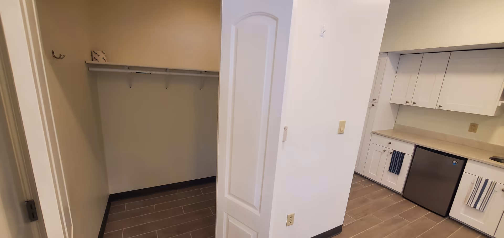 Interior view showing a small walk-in closet with a shelf and hooks on the left side, and a kitchen area with white cabinets, a small refrigerator, and a countertop on the right side. The floor is tiled with brown rectangular tiles.