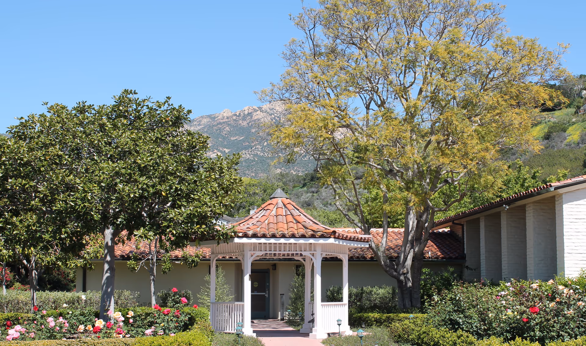 A white gazebo with a red tiled roof is centered in a garden area with blooming flowers and green bushes. Trees with green and yellow leaves surround the gazebo. Behind the gazebo is a building with a red tiled roof and light-colored walls. Mountains and a clear blue sky are visible in the background.