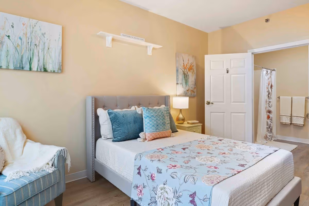 A cozy bedroom with a bed featuring a tufted gray headboard, white bedding, and decorative pillows in blue and peach tones. A floral-patterned throw blanket is draped across the foot of the bed. To the left, there is a blue striped armchair with a white throw blanket. The walls are painted beige and decorated with floral artwork. A nightstand with a lamp is beside the bed. An open door reveals a bathroom with a shower curtain and towels hanging on a rack.