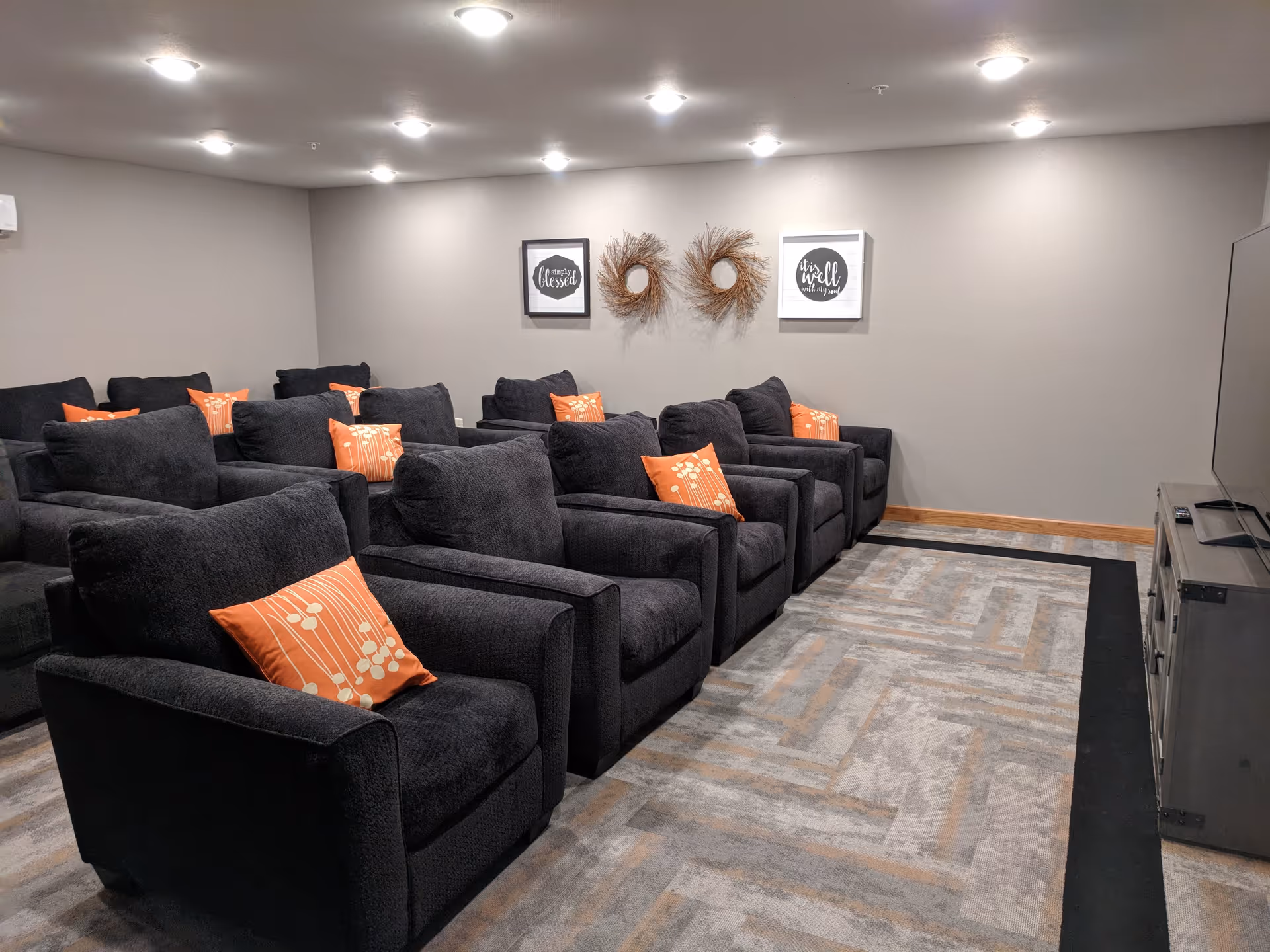 A cozy media room with multiple dark gray armchairs arranged in rows, each with an orange decorative pillow. The room has a patterned carpet, neutral-colored walls, and ceiling lights. On the wall, there are two framed quotes and two circular wreath decorations. A large flat-screen TV is positioned on a stand to the right.