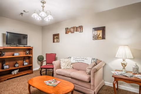 A cozy living room with a patterned sofa adorned with decorative pillows, a wooden coffee table with magazines, a red upholstered chair, a wooden shelving unit with a flat-screen TV and decorative items, a side table with a lamp and magazines, and wall art including a sign that says 'FAMILY'.