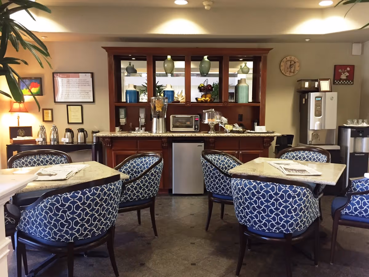 A cozy dining area with four tables surrounded by blue patterned chairs. In the background, there is a wooden cabinet with decorative vases, a blender, a toaster oven, and a fruit basket. To the right, there is a water dispenser and a coffee machine. The walls are decorated with framed pictures and a clock.