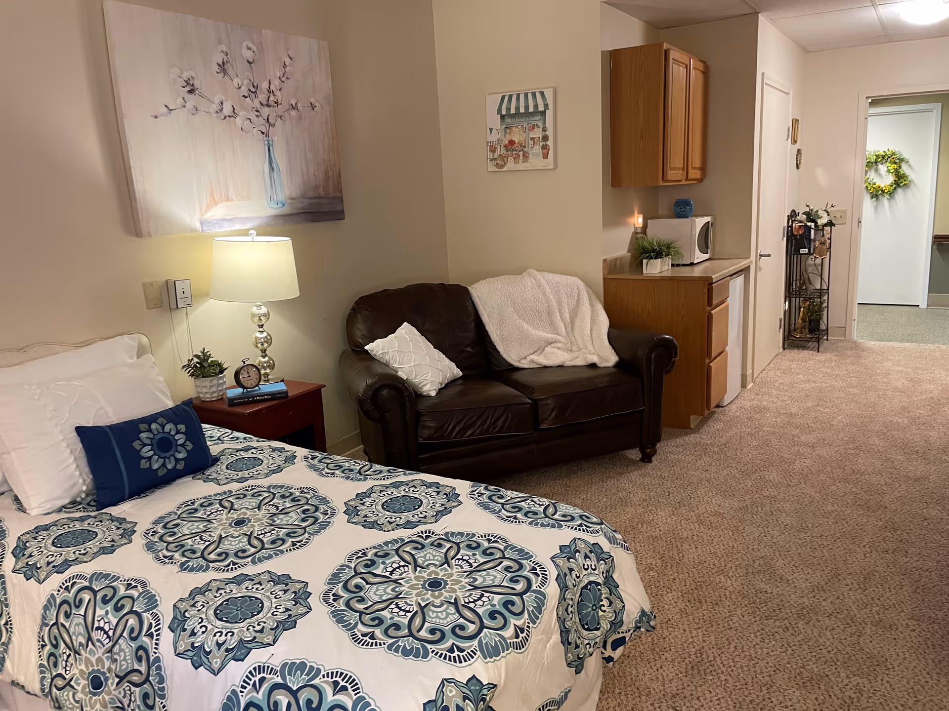 A cozy bedroom in a senior living facility featuring a bed with a blue and white patterned comforter and pillows, a bedside table with a lamp, clock, and plant, a dark brown leather loveseat with a white throw blanket and pillow, wooden cabinets with a microwave on the countertop, and a hallway leading to another room decorated with a green wreath on the door.