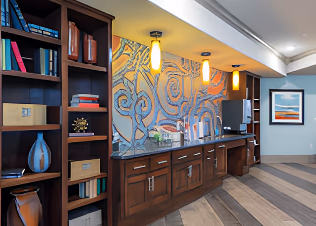 Interior view of a senior living facility featuring a wooden bookshelf filled with books and decorative items on the left, a countertop with cabinets below and three pendant lights above, and a colorful abstract mural on the wall behind the counter. A framed artwork hangs on a light blue wall in the background.