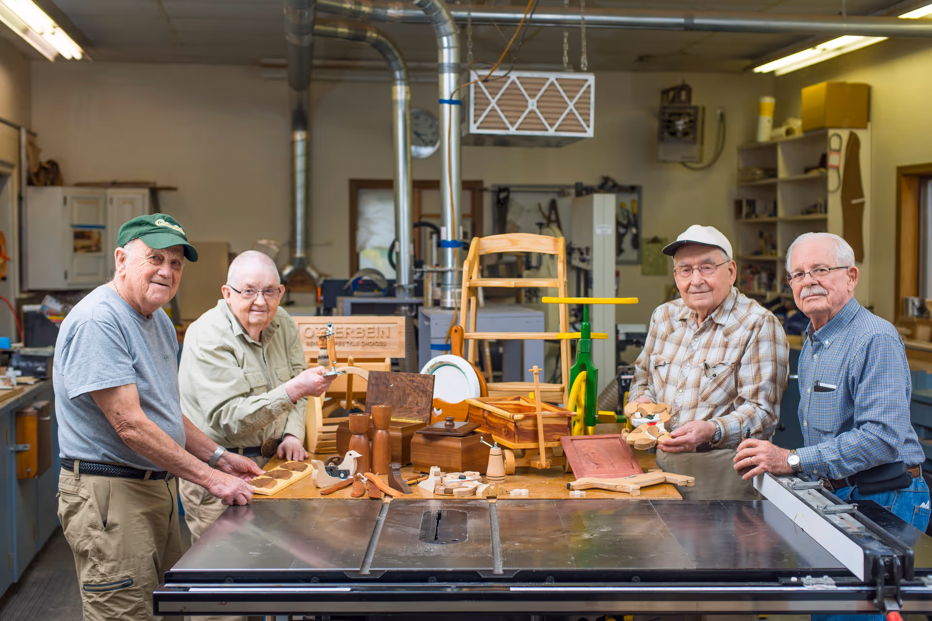 Four elderly men standing around a woodworking table in a workshop, displaying various handcrafted wooden items including a chair, toys, and decorative pieces. The workshop has tools, shelves, and ventilation ducts visible in the background.