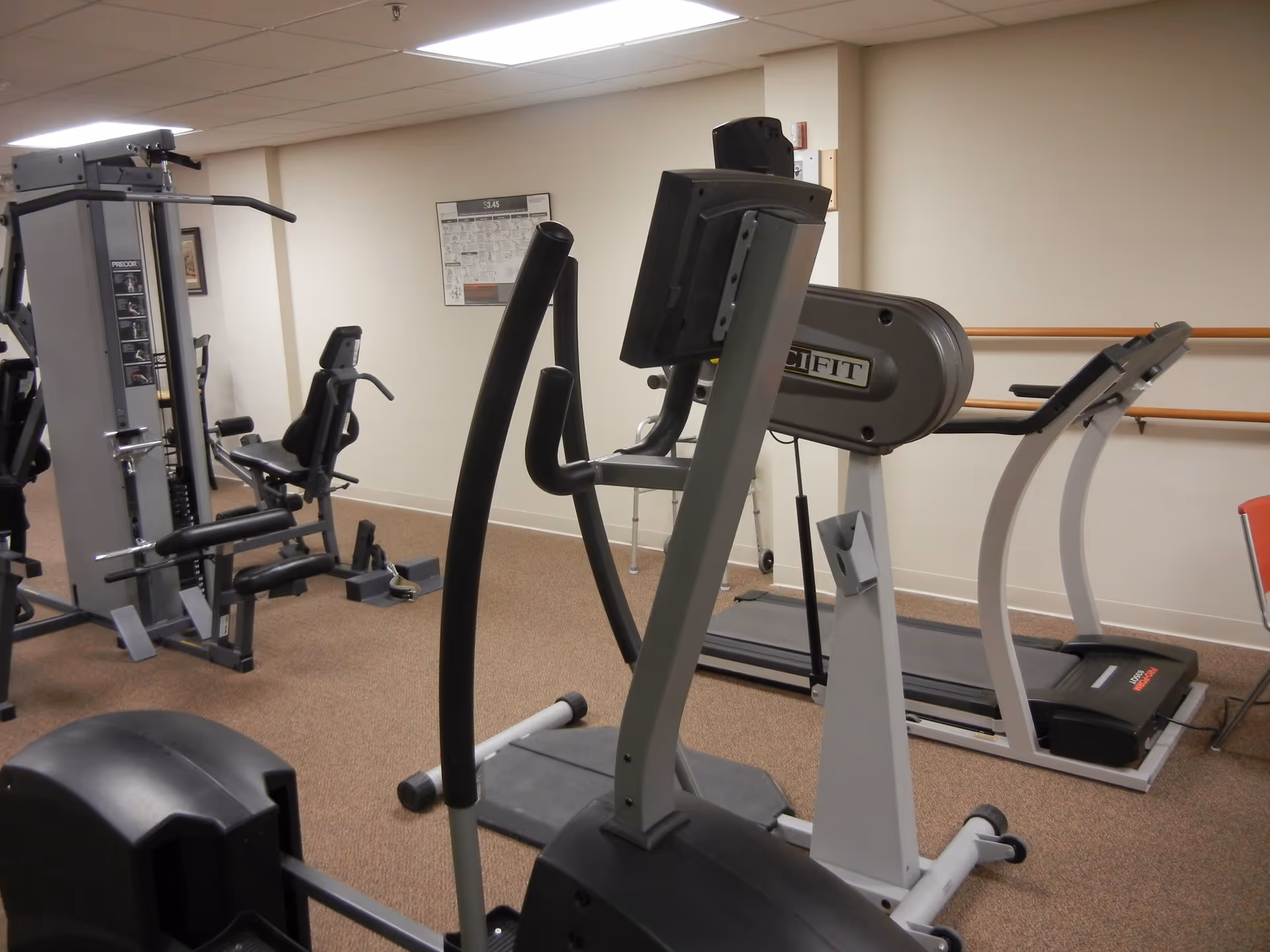 Indoor fitness room with exercise equipment including a treadmill, elliptical machine, and weight machine. The room has beige walls, carpeted floor, and handrails along one wall.