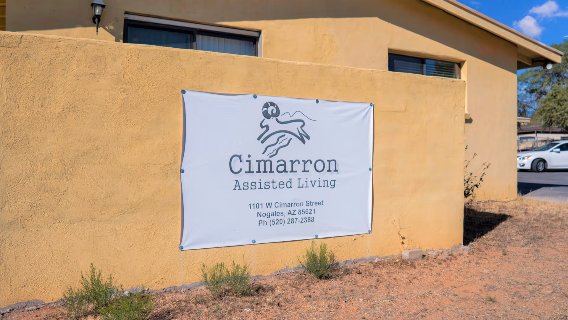 Exterior view of a beige building with a white banner sign that reads 'Cimarron Assisted Living' along with the address 1101 W Cimarron Street, Nogales, AZ 85621 and phone number (520) 287-2388. There are some small plants and dry grass in front of the building and a white car is partially visible in the background.