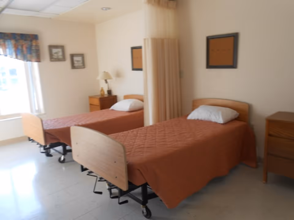 A room with two hospital-style beds with brown bedspreads and white pillows, separated by a beige privacy curtain. There is a window with a floral valance on the left wall, two framed pictures above a wooden nightstand with a lamp, and a wooden dresser on the right side of the room.