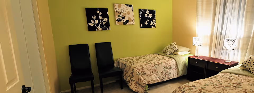 A bedroom with two single beds covered in floral bedspreads, each with pillows. Between the beds is a dark wooden nightstand with two drawers, topped with two decorative lamps. The walls are painted light green and beige, and three framed floral artworks hang above one bed. Two black chairs are placed against the green wall near the door.