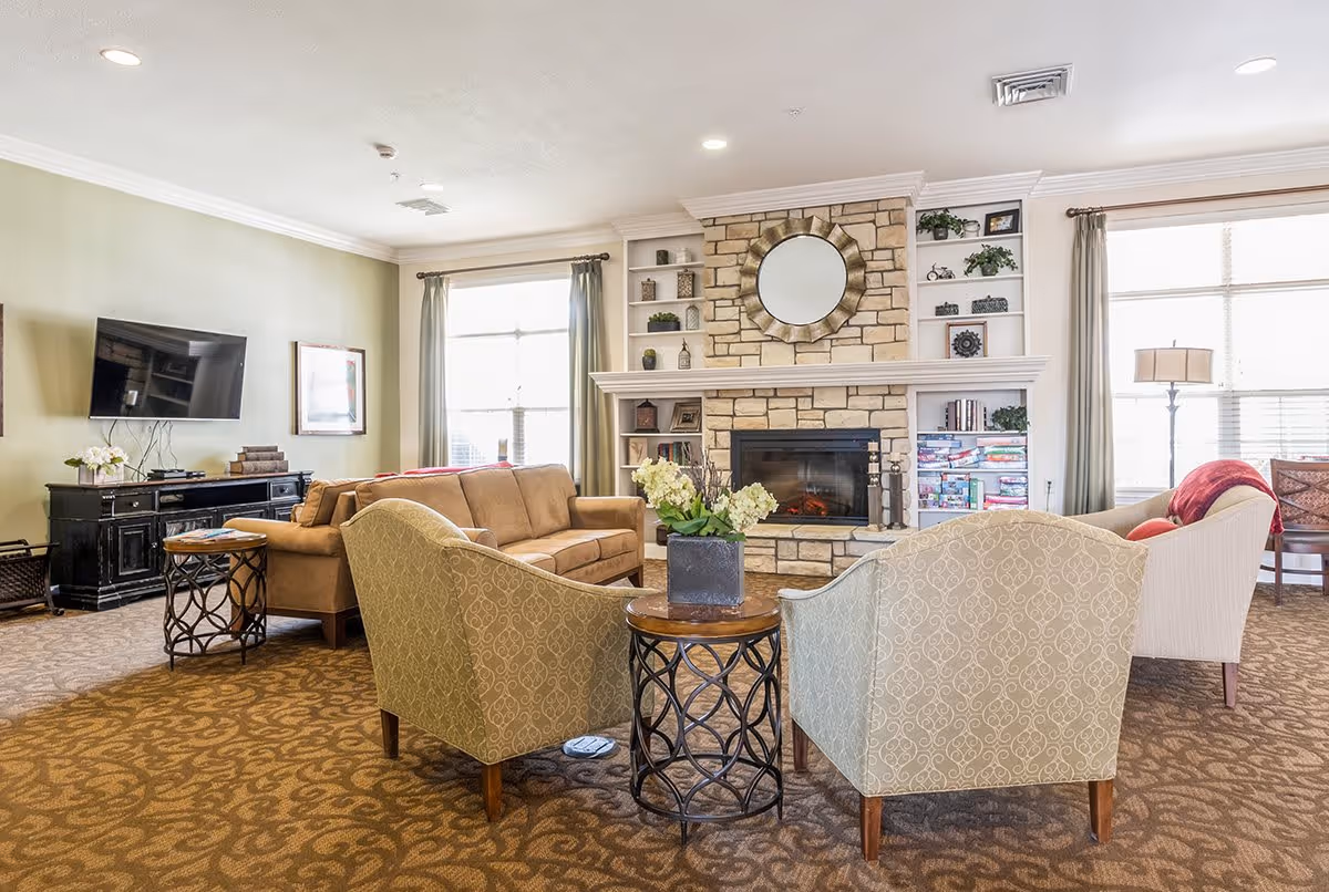 Bright, cozy common living room with sofas and armchairs arranged around a stone fireplace and wall-mounted TV.