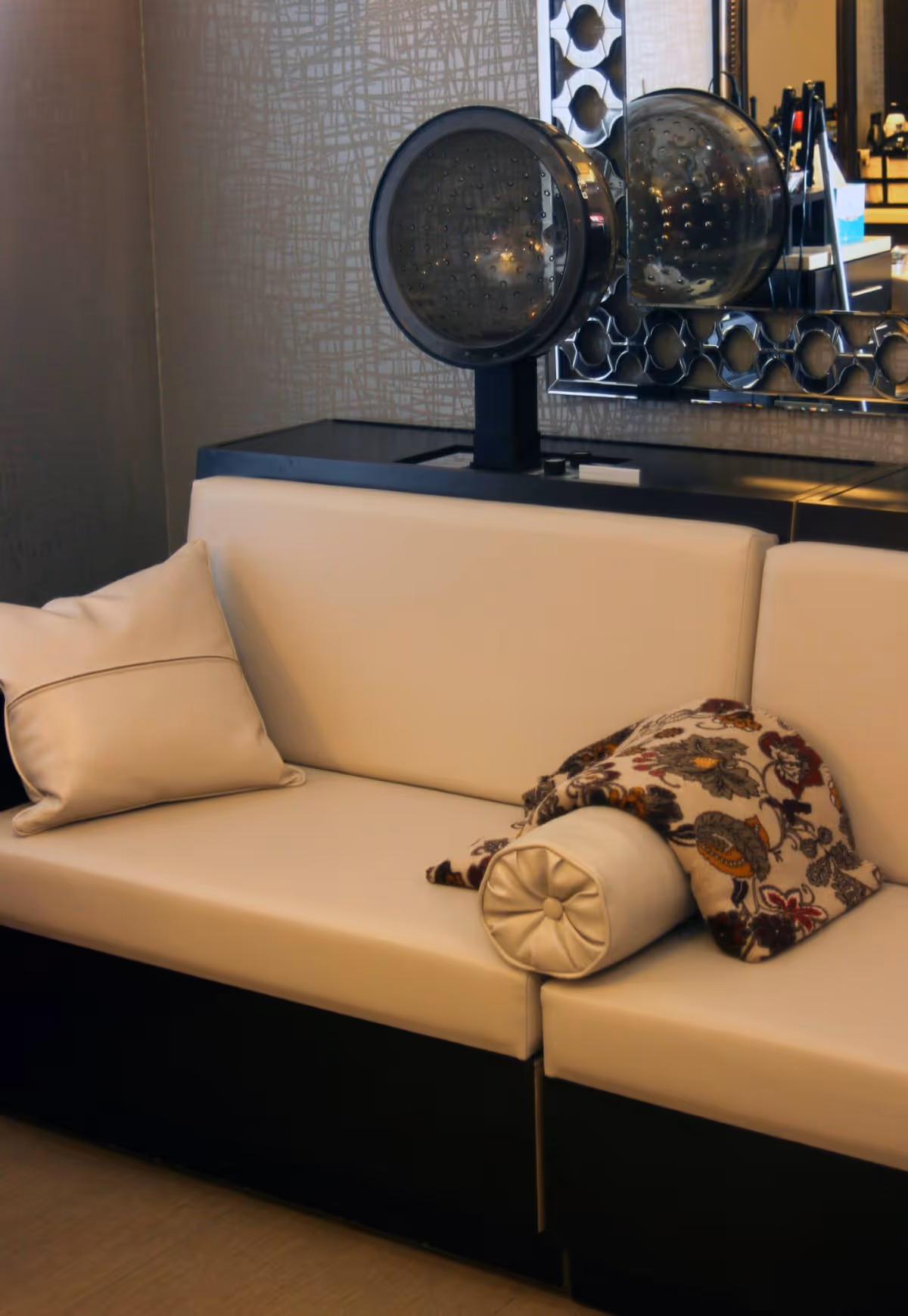 A beige cushioned bench with two pillows, one plain and one cylindrical, and a patterned throw blanket draped over the cylindrical pillow. Behind the bench is a black counter with a large round hair dryer and a decorative mirror with a geometric pattern on the frame.